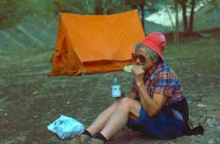 A woman enjoying a picnic in a meadow with her tent pitched behind her.